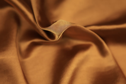 Nahaufnahme eines glänzenden Satinstoffs in edlem Kupfer-Gold mit elegantem Faltenwurf – ideal für Abendmode und Dekoration.